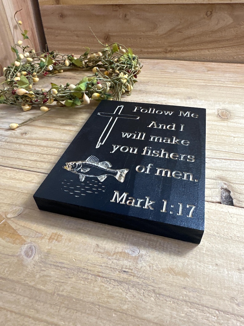 K&ouml;nnte beinhalten: Schwarze Holzplakette mit graviertem Text und Fischillustration. Der Text lautet: &bdquo;Folge mir, und ich will euch zu Menschenfischern machen. Markus 1:17.&ldquo; Ein Kreuz befindet sich &uuml;ber dem Text. Im Hintergrund befindet sich ein dekorativer Kranz.