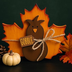 May include: A wooden Thanksgiving decoration featuring a brown turkey silhouette with an orange beak and a tag that says "GOBBLE". The turkey is set against an orange leaf-shaped background and tied with twine. A small white pumpkin is in the foreground.
