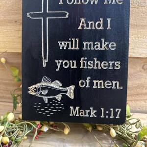 K&ouml;nnte beinhalten: Schwarze Holztafel mit graviertem Text und Bildern. Der Text lautet: &bdquo;Folge mir, und ich werde euch zu Menschenfischern machen. Markus 1:17.&ldquo; Ein Kreuz und ein Fisch sind ebenfalls auf der Tafel eingraviert. Dekorativer Artikel.