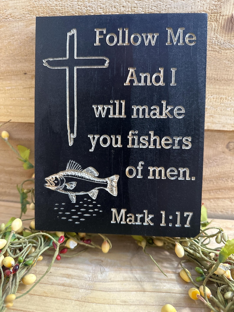 K&ouml;nnte beinhalten: Schwarze Holztafel mit graviertem Text und Bildern. Der Text lautet: &bdquo;Folge mir, und ich werde euch zu Menschenfischern machen. Markus 1:17.&ldquo; Ein Kreuz und ein Fisch sind ebenfalls auf der Tafel eingraviert. Dekorativer Artikel.