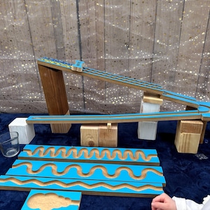 May include: Wooden marble run set with blue and natural wood accents. The set includes ramps, blocks, and curved track pieces. A child is visible in the foreground. The background has a blue surface and a wooden backdrop.
