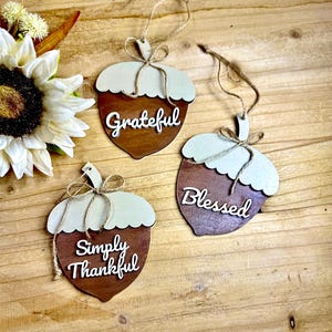 May include: Three wooden acorn ornaments with the words "Grateful", "Blessed", and "Simply Thankful" in white script. Each ornament has a light brown top and a darker brown bottom, with a twine loop for hanging. A sunflower is in the background.