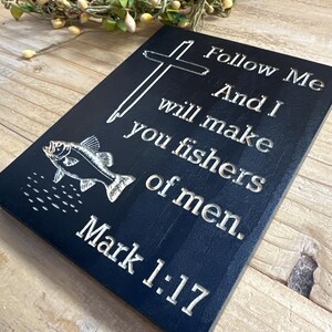 K&ouml;nnte beinhalten: Eine schwarze Holztafel mit wei&szlig;em graviertem Text und einer Fischillustration. Der Text lautet: &bdquo;Folge mir, und ich will euch zu Menschenfischern machen. Markus 1:17.&ldquo; Ein Kreuz ist ebenfalls auf der Tafel eingraviert. Ein dekorativer Kranz befindet sich im Hintergrund.