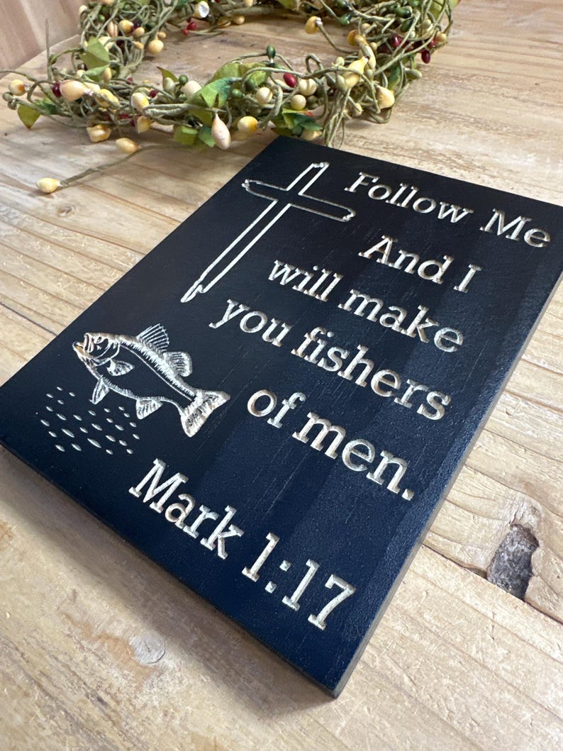 K&ouml;nnte beinhalten: Eine schwarze Holztafel mit wei&szlig;em graviertem Text und einer Fischillustration. Der Text lautet: &bdquo;Folge mir, und ich will euch zu Menschenfischern machen. Markus 1:17.&ldquo; Ein Kreuz ist ebenfalls auf der Tafel eingraviert. Ein dekorativer Kranz befindet sich im Hintergrund.