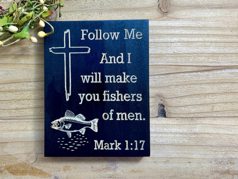 K&ouml;nnte beinhalten: Dunkelblaues Holzschild mit wei&szlig;em Text und einer Kreuz- und Fischillustration. Der Text lautet: &bdquo;Folge mir, und ich werde dich zu Menschenfischern machen. Markus 1:17.&ldquo;