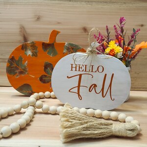 Puede incluir: Dos letreros de madera con forma de calabaza, uno naranja con un patrón de hojas y el otro blanco con el texto "Hello Fall" en letras naranjas. Un hilo de cuentas de madera y un borla marrón están en primer plano.