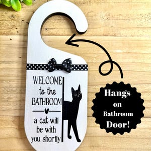 May include: White wooden bathroom door hanger with a black cat silhouette and the text "WELCOME to the BATHROOM a cat will be with you shortly." A black bow and polka dot ribbon accent the top. The sign reads "Hangs on Bathroom Door!"