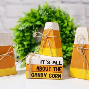 Può includere: Decorazioni in legno a forma di caramelle di mais bianche, arancioni e gialle, legate con spago. Una pila di libri recita "IT'S ALL ABOUT THE CANDY CORN". Una ghirlanda verde è sullo sfondo.