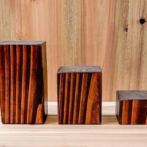 May include: Three wooden display blocks of varying sizes, all with a dark brown stain and a grooved texture. The blocks are arranged in a row from largest to smallest.