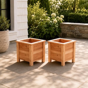 May include: Two square wooden planters with a natural light brown finish. Each planter features a sturdy frame with four corner posts, a flat top rim, and an open interior. The wood grain is visible on the panels of these outdoor containers.