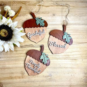 May include: Three wooden acorn decorations with the words "Grateful", "Blessed", and "Simply Thankful" in white script. Each acorn has a brown top, a light wood body, and a green leaf accent. They hang from twine.
