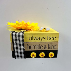 May include: A decorative wooden block set with a yellow and black color scheme. The top block is yellow with the words "always bee" and a small bee figurine. The middle block reads "humble & kind". A yellow flower and black and white ribbon are also present.