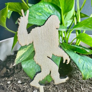 Puede incluir: Una estaca de planta de madera de Bigfoot con un gesto de signo de la paz. El recorte de madera de color claro tiene un contorno oscuro y se inserta en el suelo con plantas de hojas verdes en el fondo. Bigfoot está haciendo el signo de la paz.