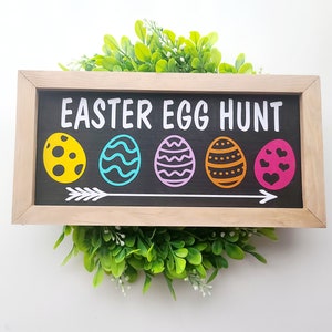 Może przedstawiać: Drewniany znak z napisem "Easter Egg Hunt" białymi literami. Znak ma pięć kolorowych jajek z różnymi wzorami i strzałkę wskazującą w prawo.