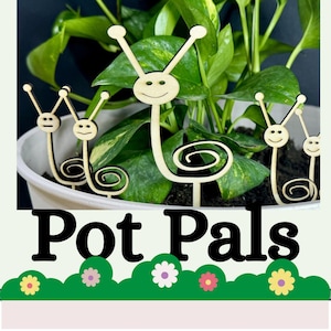 May include: Wooden plant stakes with smiling face designs, labeled "Pot Pals", are placed in a white pot with green leafy plants. The stakes have a whimsical, cartoonish appearance, adding a decorative touch to indoor plants.