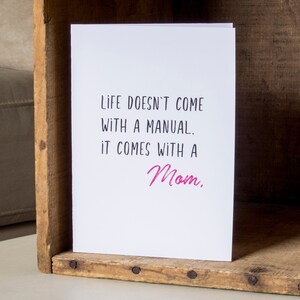 May include: White card with black text that reads "Life doesn't come with a manual. It comes with a Mom." The word "Mom" is written in pink.