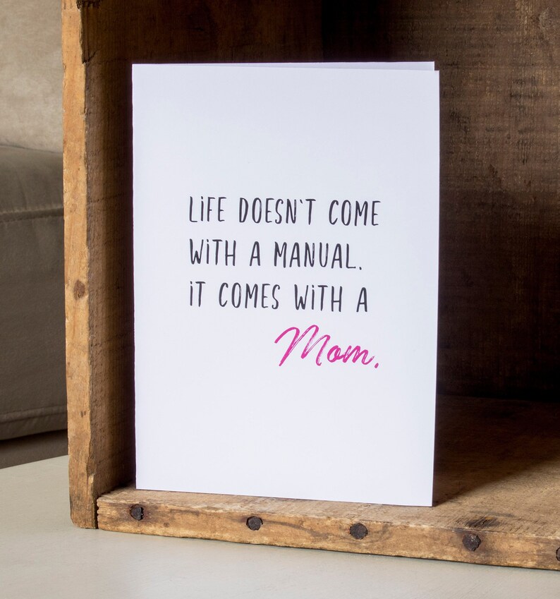 May include: White card with black text that reads "Life doesn't come with a manual. It comes with a Mom." The word "Mom" is written in pink.