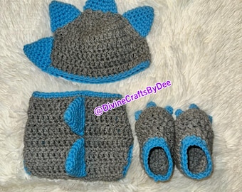 May include: A gray and blue crocheted dinosaur costume set for a baby. The set includes a hat, booties, and a diaper cover. The hat has a pointed top with spikes and the booties have three toes. The diaper cover has a tail and a dinosaur face on the front.