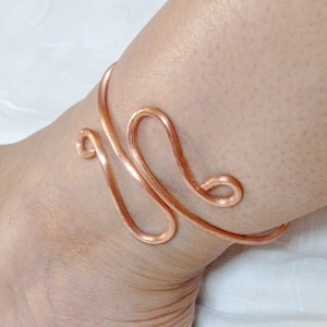 May include: A copper ankle bracelet with a swirling design. The bracelet is made of a single piece of wire that is twisted and shaped into a unique pattern.