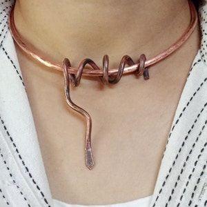 May include: Copper-coloured necklace with a choker-style band, a spiralled design, and a dangling, pointed pendant. The necklace is worn against a white and black patterned shirt.