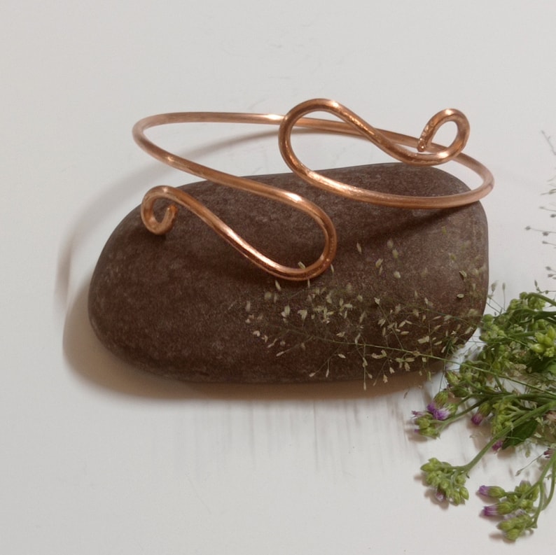May include: A copper wire bracelet with a simple, elegant design. The bracelet is made of a single piece of wire that is twisted and shaped into a delicate, flowing pattern.