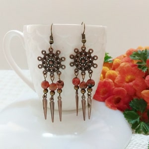 Pode incluir: Um par de brincos pendurados de cor cobre. Cada brinco apresenta um topo decorativo vazado em forma de estrela, com contas vermelhas e castanhas e três pingentes pontiagudos de cor cobre. Os brincos são exibidos em uma superfície branca.