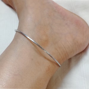 Puede incluir: Un brazalete de tobillo plateado con un diseño simple y delgado.