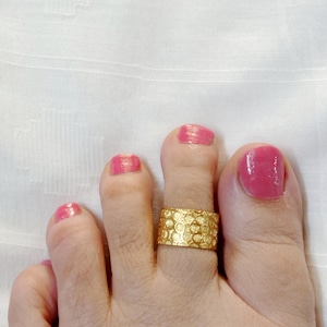 May include: Gold toe ring with a textured design. The ring is worn on the second toe of a foot with pink nail polish.