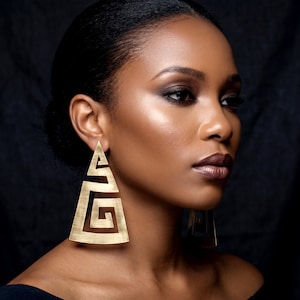 May include: Gold-coloured, geometric earrings with a triangular shape and a cut-out design. The earrings feature a spiral pattern within the triangle. The earrings are worn by a person with dark skin and dark hair. The background is dark.