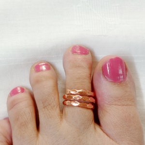 Puede incluir: Primer plano de un pie con tres anillos de dedo del pie de cobre apilados. El dedo del pie está pintado con esmalte de uñas rosa.