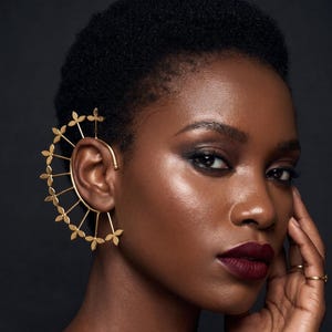 May include: A gold-colored ear cuff featuring a floral design. The ear cuff curves around the ear and is adorned with small, delicate flower-shaped accents. The jewelry piece is worn on a person with dark skin and short, dark hair.