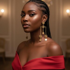 May include: A woman wearing gold-coloured, dangling earrings with floral and geometric shapes. The earrings are suspended from chains. She is wearing a red off-the-shoulder dress. Her hair is styled in braids.