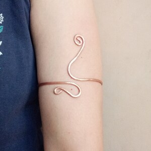 Op de afbeelding: Koperen armmanchet met een wervelend ontwerp. De manchet wikkelt zich om de bovenarm met een horizontale band en decoratieve gebogen accenten. De sieraden hebben een warme, roodbruine kleur.