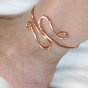May include: A copper wire ankle bracelet with a unique, abstract design. The bracelet is made of a single piece of wire that is twisted and shaped into a decorative pattern.