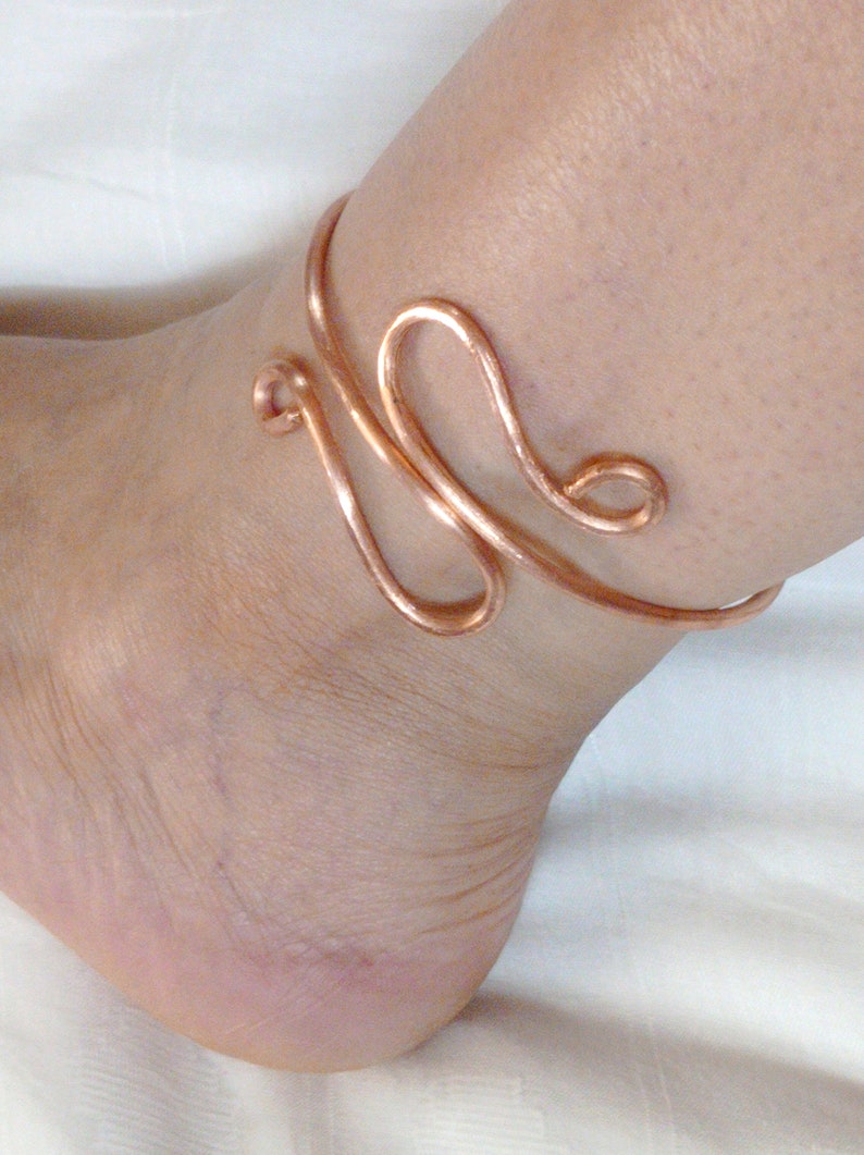May include: A copper wire ankle bracelet with a unique, abstract design. The bracelet is made of a single piece of wire that is twisted and shaped into a decorative pattern.