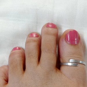 May include: A silver toe ring with a simple, straight design. The ring is worn on the big toe of a foot with pink nail polish.