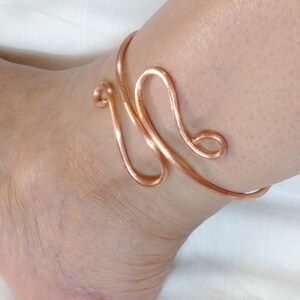 May include: A copper wire ankle bracelet with a unique, twisted design. The bracelet is made of a single piece of wire that is bent and shaped into a decorative pattern.