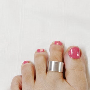 Pode incluir: Um anel de dedo do pé de prata com uma banda larga em um pé com esmalte de unha rosa.