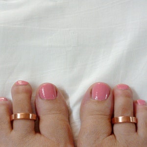 Puede incluir: Dos anillos de dedo del pie de cobre en un par de pies con esmalte de uñas rosa.