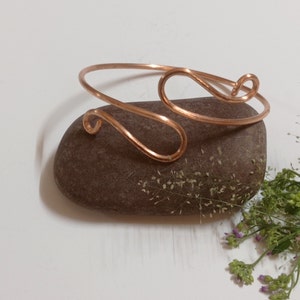 May include: A simple copper wire bracelet with a unique, curved design. The bracelet is shown on a brown rock with green foliage.