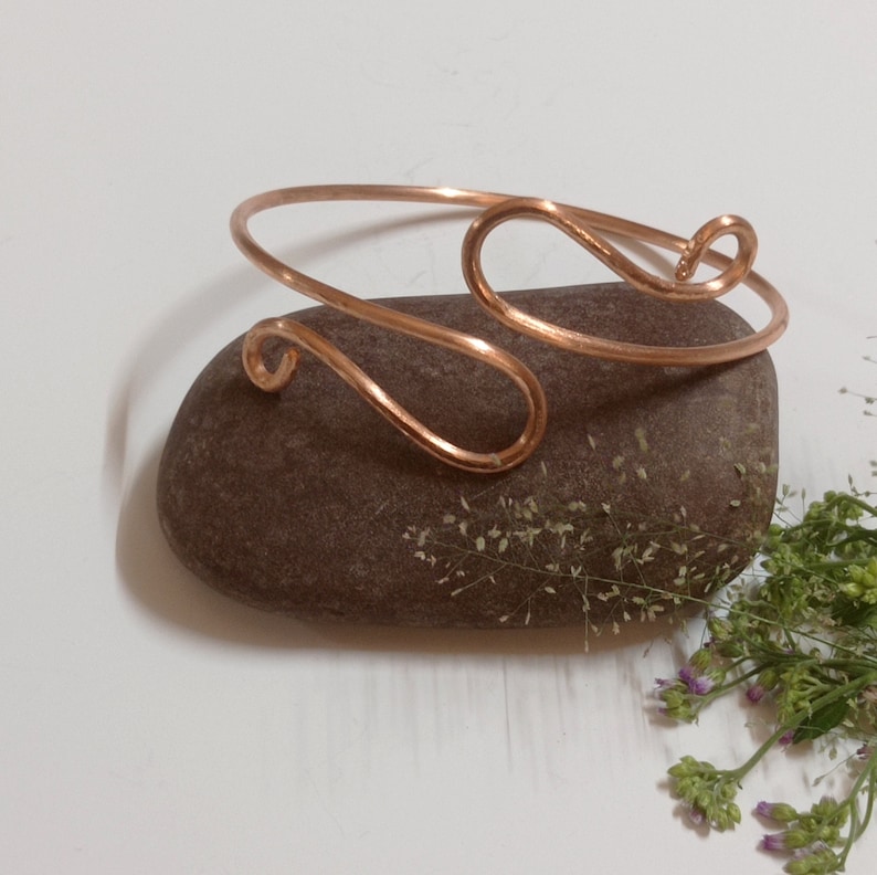 May include: A simple copper wire bracelet with a unique, curved design. The bracelet is shown on a brown rock with green foliage.