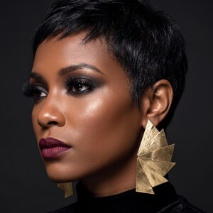 May include: A woman with short black hair and striking eye makeup. She wears large, gold geometric earrings. Her lips are a deep burgundy colour, and she is wearing a black turtleneck. The background is a dark grey.
