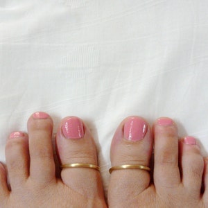 May include: Close-up of two feet with pink nail polish and gold toe rings. The feet are resting on a white surface. The toe rings are simple, thin bands of gold. The nail polish is a soft, rosy pink color.