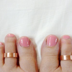 Puede incluir: Dos dedos del pie con esmalte de uñas rosa y anillos de dedo del pie de cobre.