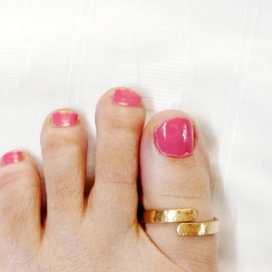 May include: A gold toe ring with a hammered finish. The ring is worn on the big toe of a foot with pink nail polish.