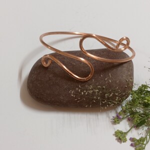May include: A simple copper wire bracelet with a unique, curved design. The bracelet is shown on a brown rock with green foliage.