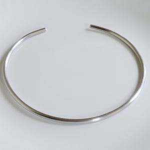 May include: A silver-toned metal choker necklace featuring a smooth, rounded, and continuous circular band design with a small open gap at the top. The piece has a polished, reflective surface and a simple, unadorned, and uniform cylindrical shape.