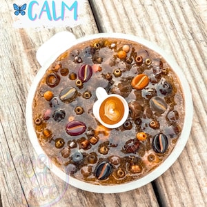 May include: A round, coffee-themed resin art piece with a white border. The artwork features coffee beans, beads, and a miniature coffee cup with a heart-shaped design. The word "CALM" is written in light blue with a butterfly graphic.