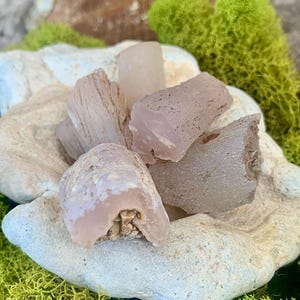 May include: A collection of five pink and white agate stones with a rough, natural texture. The stones are arranged on a white, textured surface.