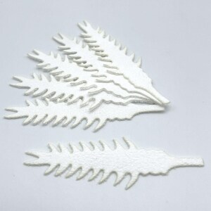 May include: Several white, feather-shaped craft embellishments are arranged on a white surface. The feathers have a textured appearance and a jagged edge, resembling a stylized leaf or plant element. The image is well-lit, highlighting the details of the craft supplies.